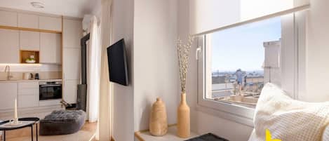 Ein modernes, kompaktes Wohnzimmer mit Küchenzeile, einem Flachbildfernseher, einer gemütlichen Sitzecke mit Blick auf die Stadt und einem Vorhang vor dem Fenster.