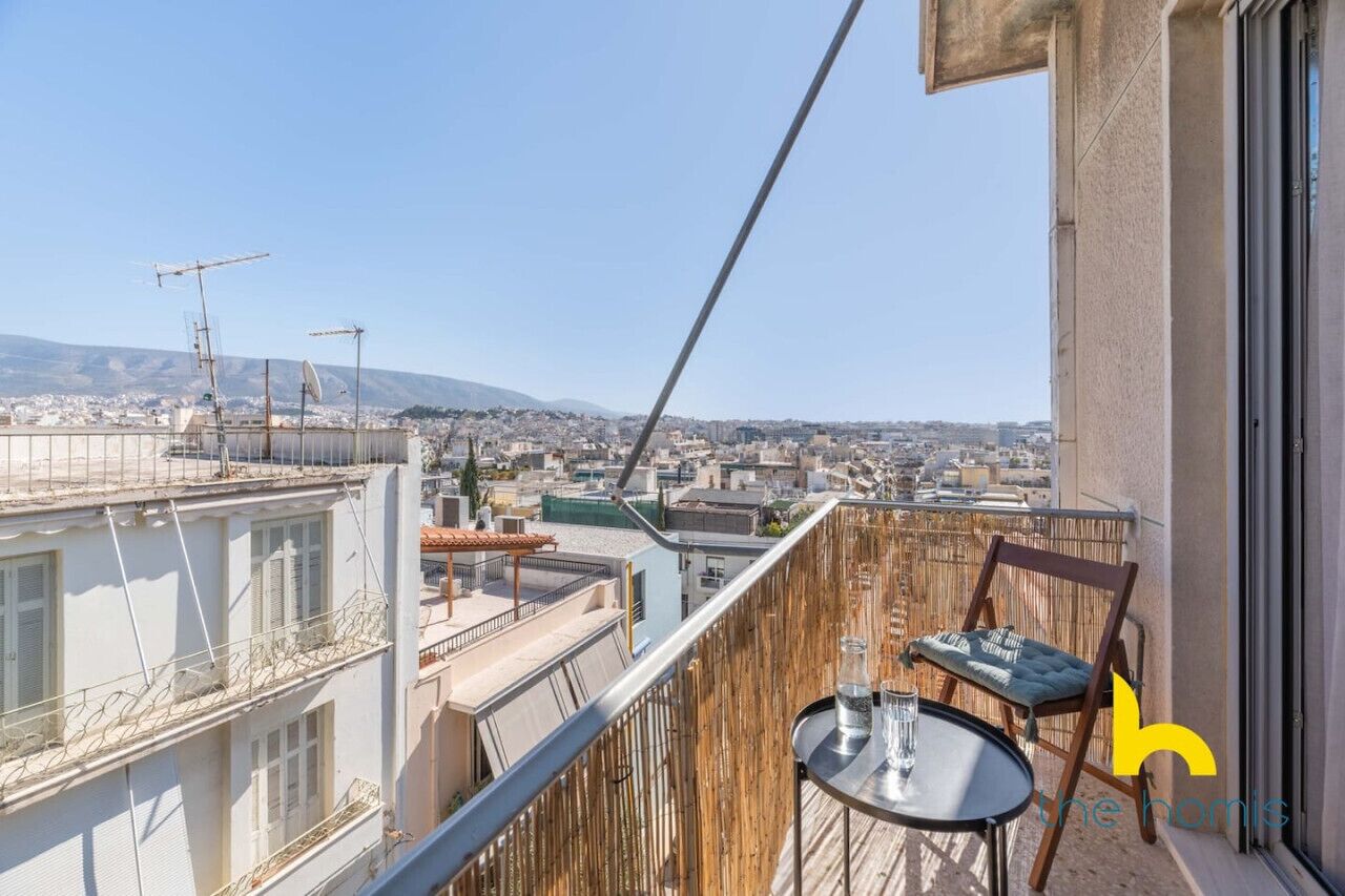 Un balcon avec une table et des chaises, d’où l’on peut admirer un panorama urbain avec des bâtiments et des montagnes à l’horizon.