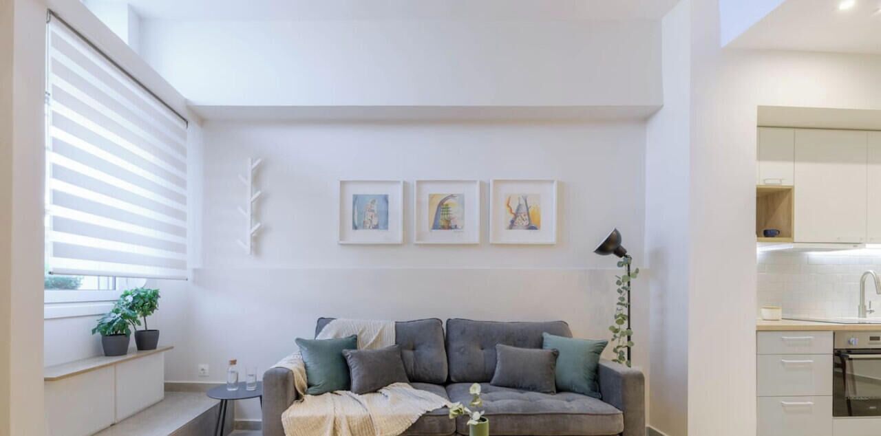 Una sala de estar moderna con un sofá gris, una mesita auxiliar y una planta en maceta. Hay una ventana con persianas, una zona de cocina con armarios y tres cuadros en la pared.