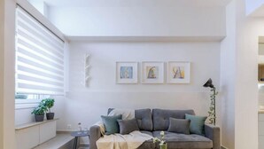 A modern living room with a grey sofa, a small side table, and a potted plant. There is a window with blinds, a kitchen area with cabinets, and three framed artworks on the wall. - Cozy Hub - Work, Relax, Explore - Prime Location (Athina)