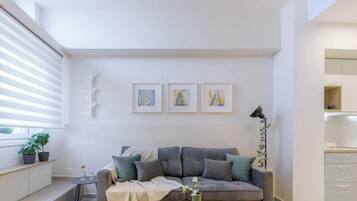 Una sala de estar moderna con un sofá gris, una mesita auxiliar y una planta en maceta. Hay una ventana con persianas, una zona de cocina con armarios y tres cuadros en la pared.