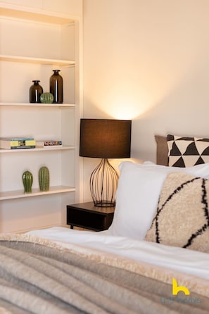 Ein ordentlich eingerichtetes Schlafzimmer mit einem Bett, einem Nachttisch mit Lampe, einem Bücherregal und dekorativen Kissen.