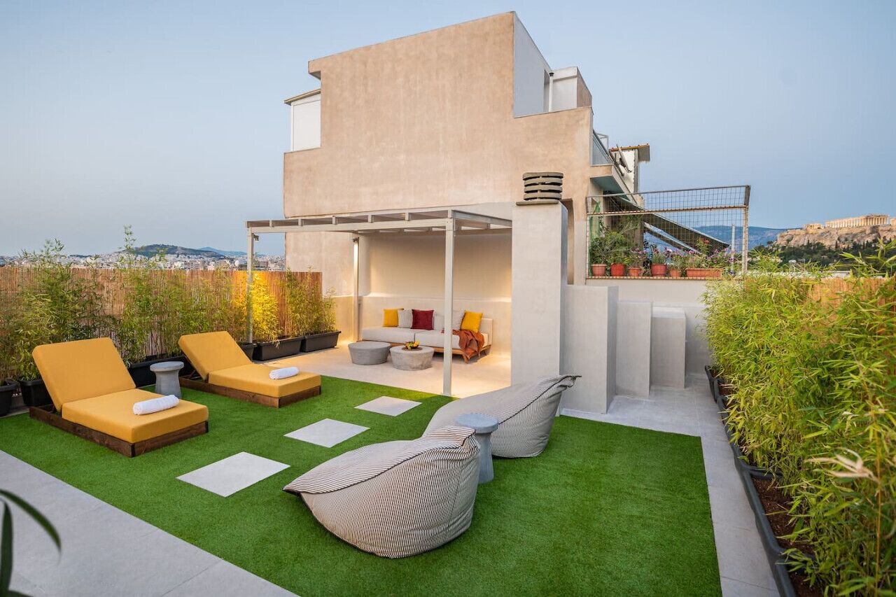 Ein moderner Außenbereich mit einer Sofagarnitur, zwei Lounge-Stühlen und einem Sitzsack auf Kunstrasen, mit Blick auf eine Stadtlandschaft und einen Hügel.