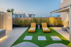 Una terraza en la azotea con dos tumbonas, césped artificial, plantas en macetas y vistas a la ciudad.