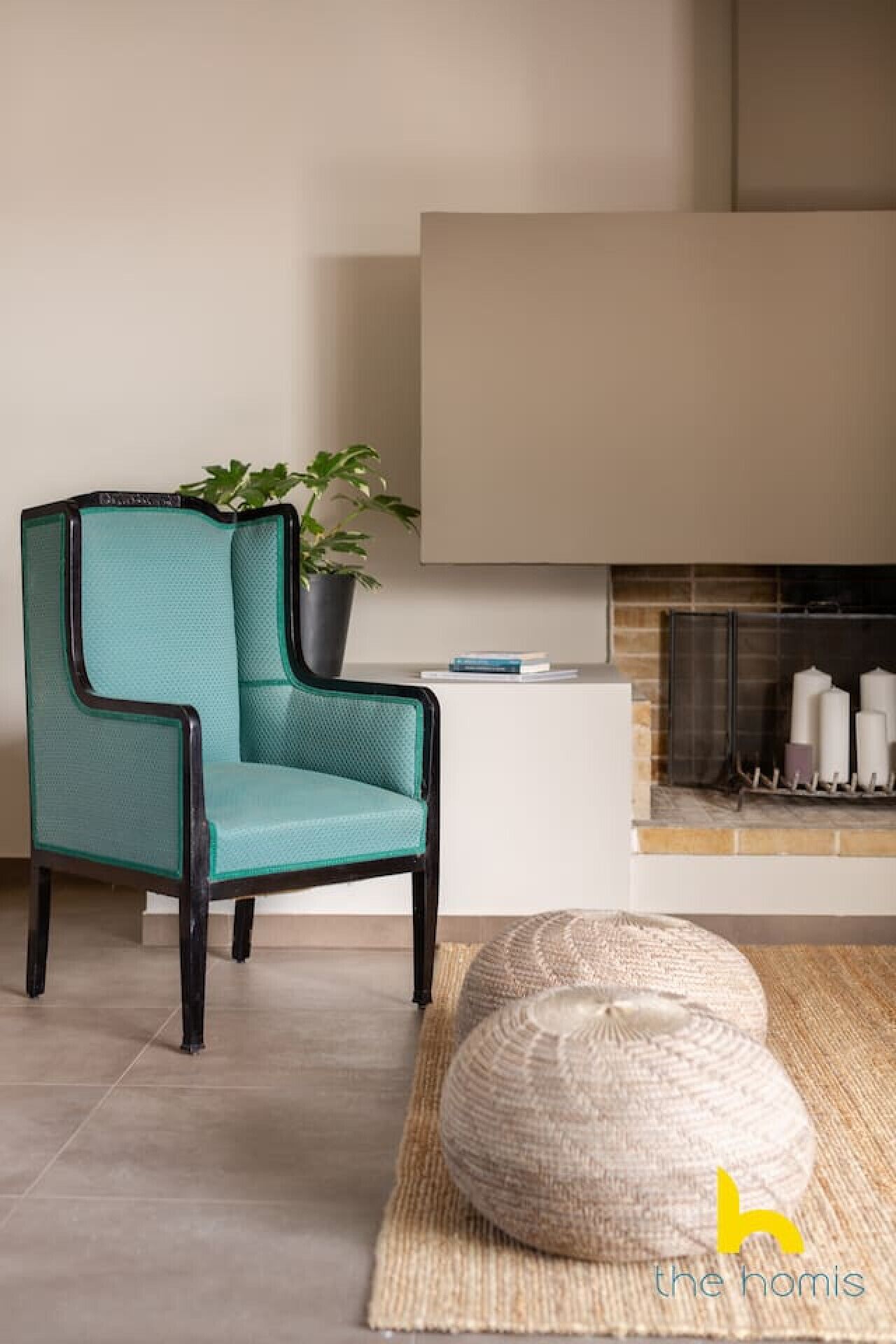 Un fauteuil de couleur turquoise avec une structure noire, une plante en pot, une petite table d’appoint blanche, une cheminée avec des bougies et un pouf tressé.