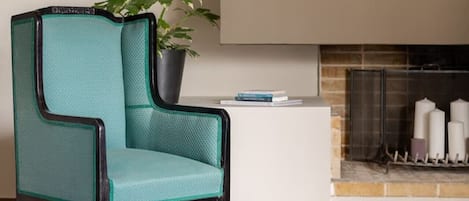 Un fauteuil de couleur turquoise avec une structure noire, une plante en pot, une petite table d’appoint blanche, une cheminée avec des bougies et un pouf tressé.