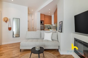 A modern living room with a grey sofa, a black coffee table, and a flat-screen TV. There is a kitchen in the background with wooden cabinets and a tiled backsplash. - Enigmatic Romantic Nest w/Fireplace Near Acropolis (Athina)