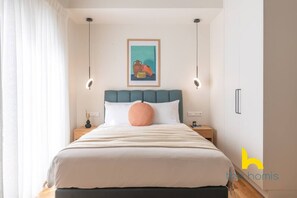 A neatly arranged bedroom with a bed, a bedside table, a nightstand, a wall-mounted artwork, and a window with curtains. - Enigmatic Romantic Nest w/Fireplace Near Acropolis (Athina)