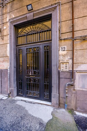 Entrada de um edifício com uma grande porta preta ornamentada, uma pequena janela acima da porta e o número '12' na parede.