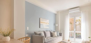A modern living room with a light blue wall, a grey sofa, a wooden chair, and a large window with a view of the outside. - Koukaki Elegance Athenian Retreat near Acropolis! (Athina)