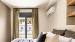 A modern hotel room with a bed, curtains, an air conditioning unit, and a view of buildings outside. - Premium Retreat w/ Patio, Parking & Acropolis View (Athina)