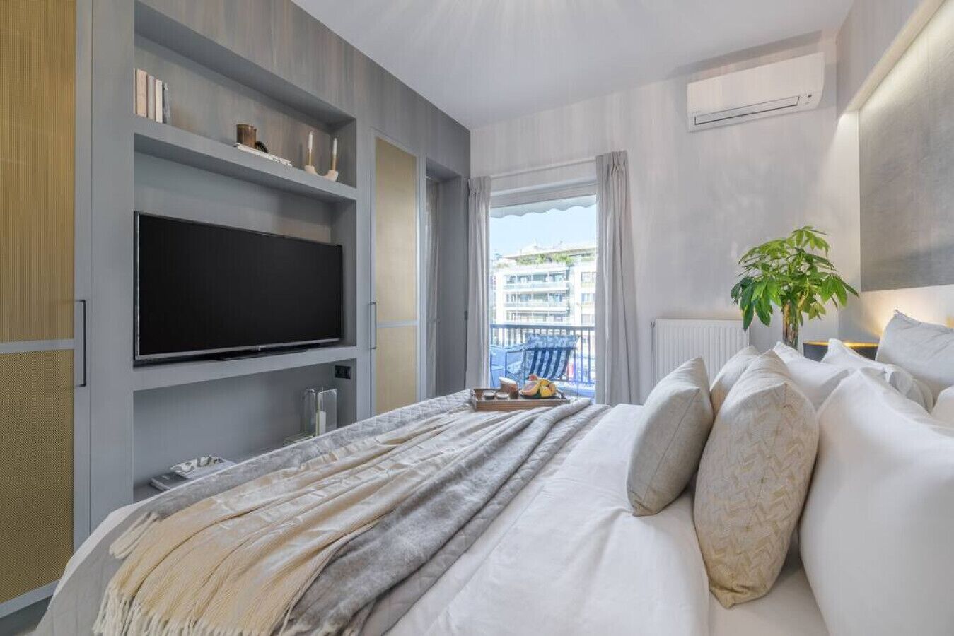 A modern hotel room with a large bed, a flat-screen TV mounted on the wall, a balcony with a view of buildings, and a potted plant.