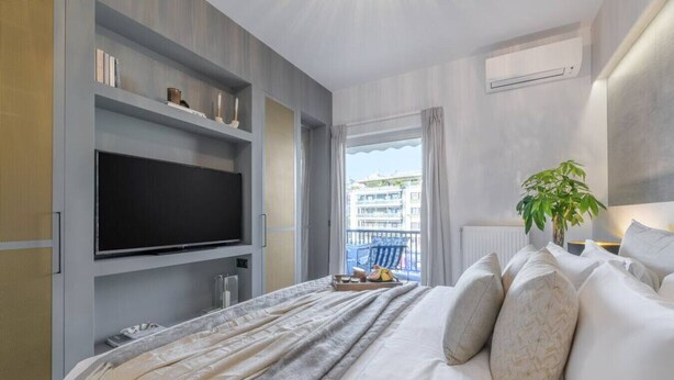 A modern hotel room with a large bed, a flat-screen TV mounted on the wall, a balcony with a view of buildings, and a potted plant. - 50 Shades of Athens, Kingsize Suite with Jacuzzi! (Athina)