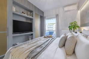 A modern hotel room with a large bed, a flat-screen TV mounted on the wall, a balcony with a view of buildings, and a potted plant. - 50 Shades of Athens, Kingsize suite with Jacuzzi! (Athina)