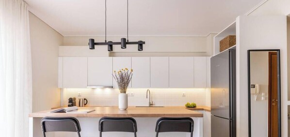 Une cuisine moderne avec des armoires blanches, un plan de travail en bois, un luminaire noir suspendu et un réfrigérateur en acier inoxydable.