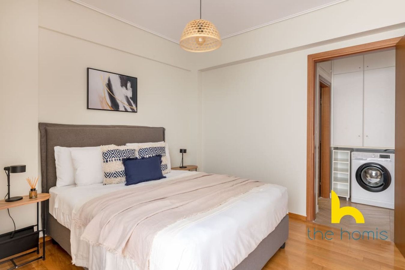 A bedroom with a bed, a nightstand, a lamp, a painting on the wall, and a washing machine in the adjacent room.