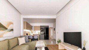 Una sala de estar moderna con sofá, una mesa de centro y un televisor de pantalla plana. En el fondo, hay una cocina con un refrigerador y un microondas.