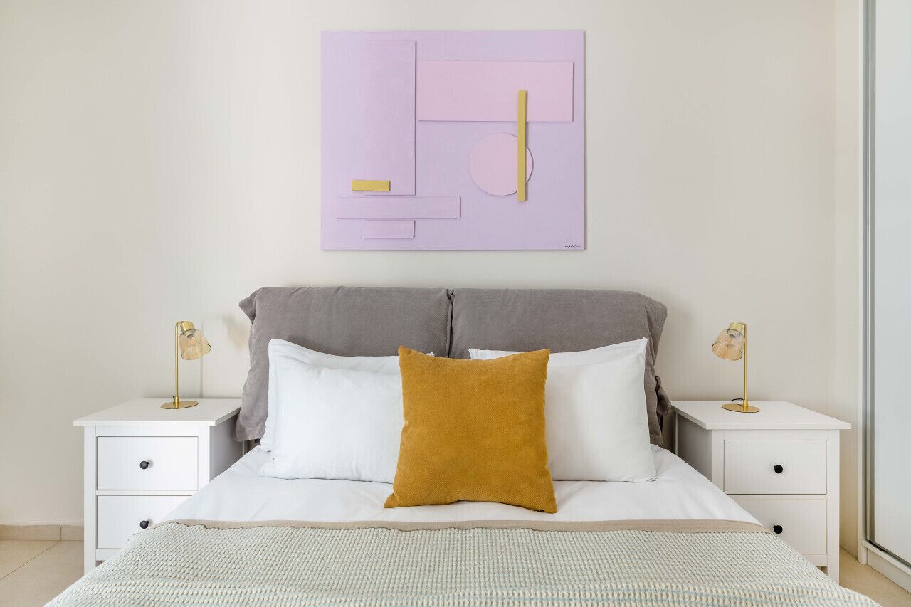 A bedroom with a bed, two bedside tables, a nightstand, and a wall-mounted abstract artwork.