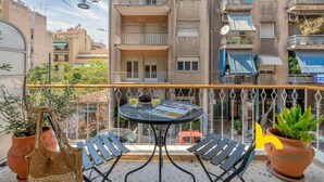 Un balcó amb taula i cadires, plantes en testos i vista d'un edifici de diversos pisos.