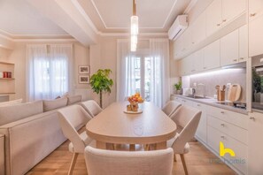 Eine moderne Küche mit weißen Schränken, ein Essbereich mit rundem Tisch und Stühlen sowie ein Wohnbereich mit einer Couch und einer Pflanze.