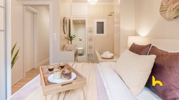 Una habitación de hotel moderna con una cama, una mesita de noche con una bandeja de pasteles, un espejo, una planta y un baño visible a través de una puerta abierta.