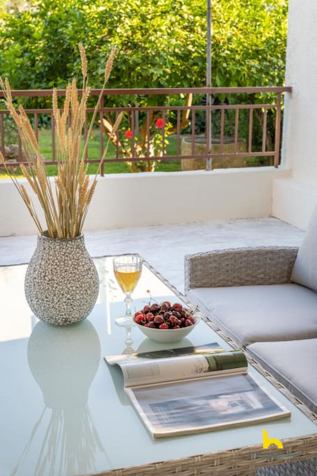 Un balcon avec une table en verre, un vase contenant des plantes séchées, un bol rempli de baies, un verre de vin et un magazine.