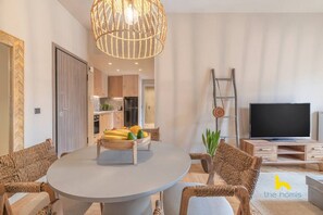 Un salon moderne avec une table ronde, des chaises en osier, un téléviseur à écran plat et un coin repas agrémenté d’un panier de fruits.