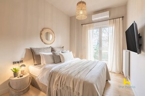 A bedroom with a bed, bedside table, lamp, and a TV mounted on the wall. - Sun-Kissed Boho Haven with Whirlpool Bliss & Patio (Athina)