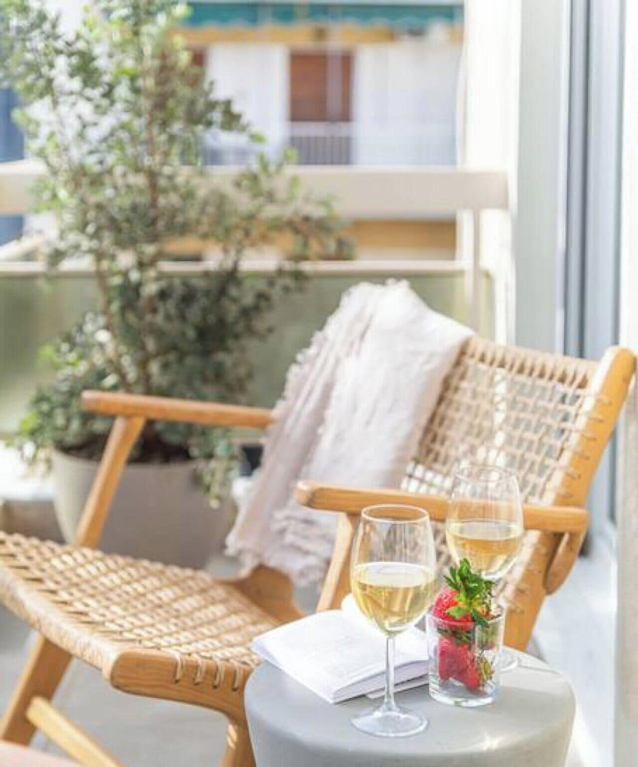 A wicker chair with a white towel, a glass of white wine, and a glass of water on a small table, with a balcony and a building in the background.