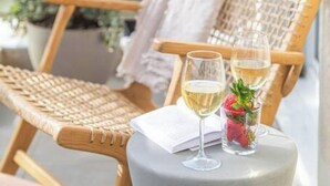 En flätad stol med en vit handduk, ett glas vitt vin och ett glas vatten på ett litet bord, med en balkong och en byggnad i bakgrunden.