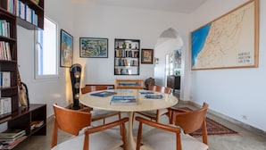 A dining area with a round table, six wooden chairs, and books on the table. There are bookshelves on the left, a map on the wall, and a lamp in the center. - Auberge Azul ifni (Sidi Ifni)