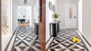 Un pasillo con piso de diseño geométrico, una puerta de madera y un espejo redondo sobre un pedestal negro.