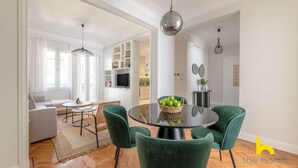 Una sala moderna con una mesa redonda de vidrio, sillas verdes, un sofá y un televisor sobre un mueble blanco.