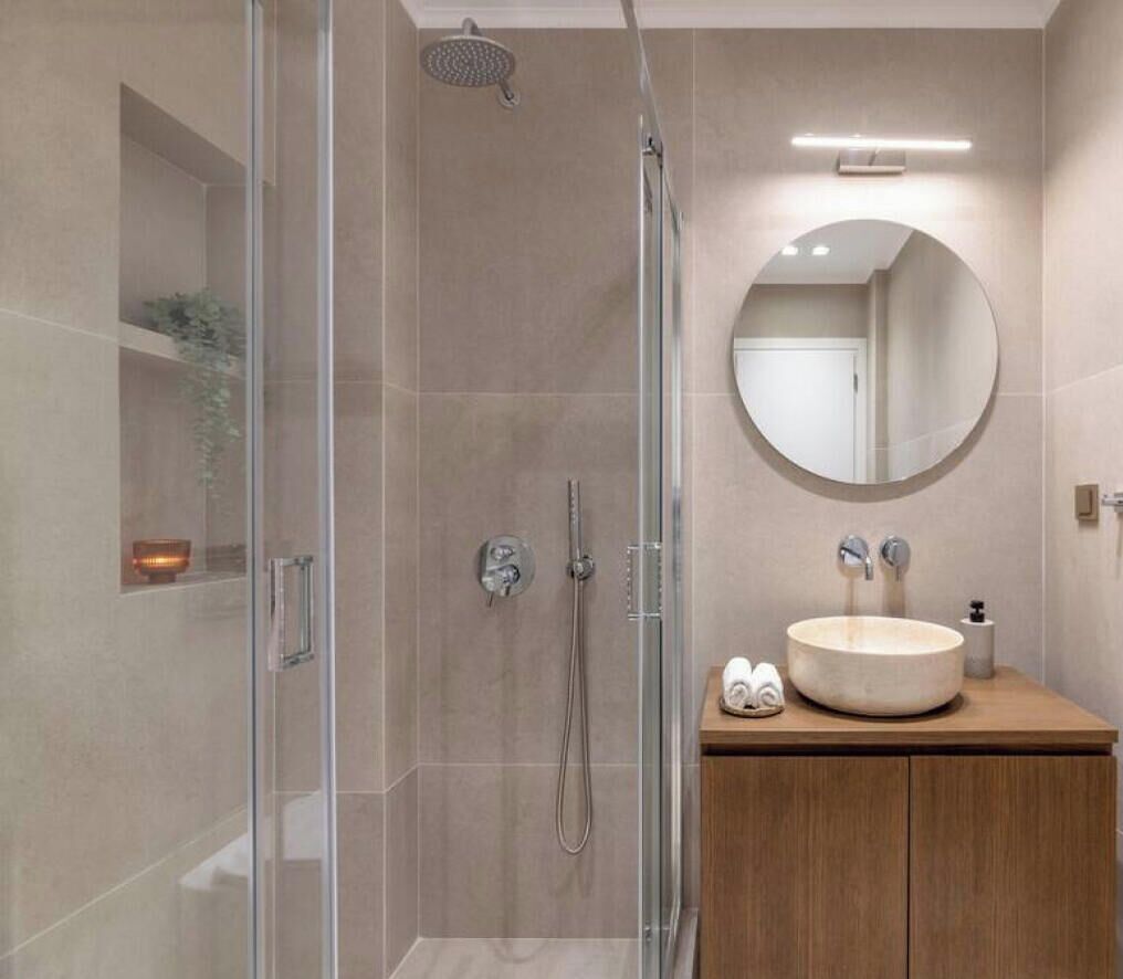A modern bathroom with a glass-enclosed shower, a round mirror, and a wooden vanity with a sink.