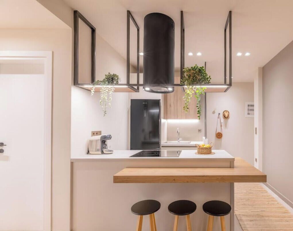 A modern kitchen with a central island, black stools, and hanging plants.