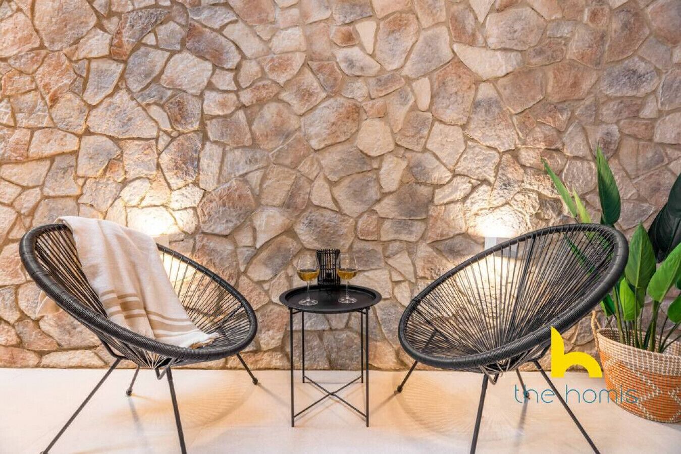 Un mur de pierres, deux fauteuils en osier, une petite table avec une lampe et une plante en pot.