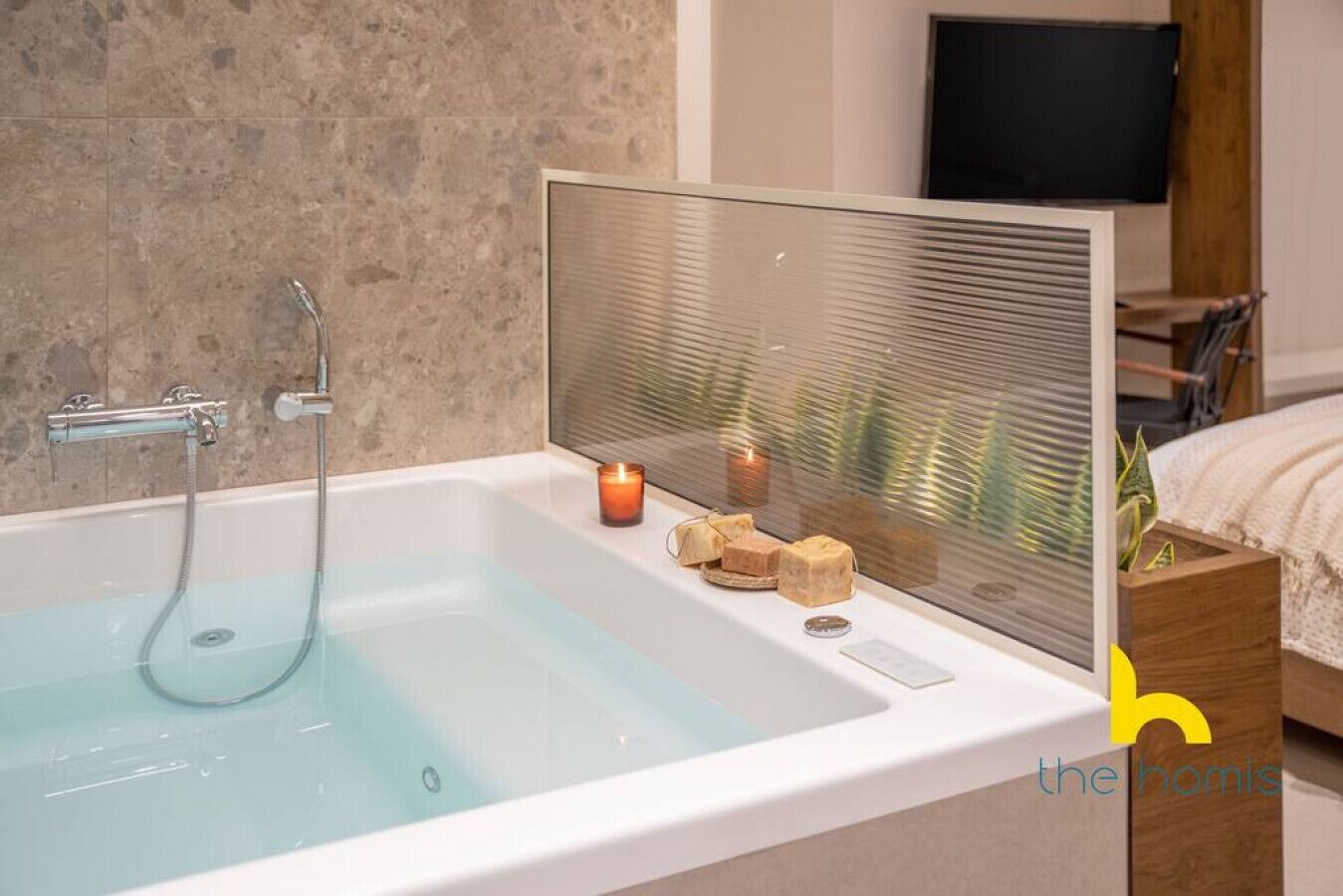A modern bathroom with a glass-enclosed bathtub, a wooden nightstand with a candle, and a TV in the background.