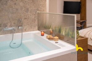 Une salle de bain moderne avec une baignoire entourée de parois en verre, une table de chevet en bois sur laquelle est posée une bougie, et un téléviseur en arrière-plan.