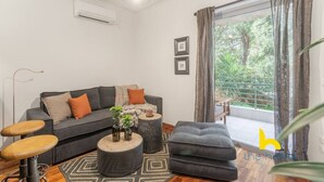A modern living room with a grey sofa, a round coffee table, and a grey ottoman. There is a window with curtains, a plant, and a picture frame on the wall. - Riviera Haven - Serene Sunny Retreat Near Seaside! (Vouliagmeni)