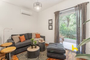 Ein modernes Wohnzimmer mit einem grauen Sofa, einem runden Couchtisch und einem grauen Hocker. Ein Fenster mit Vorhängen, eine Pflanze und ein Bilderrahmen sind an der Wand zu sehen.