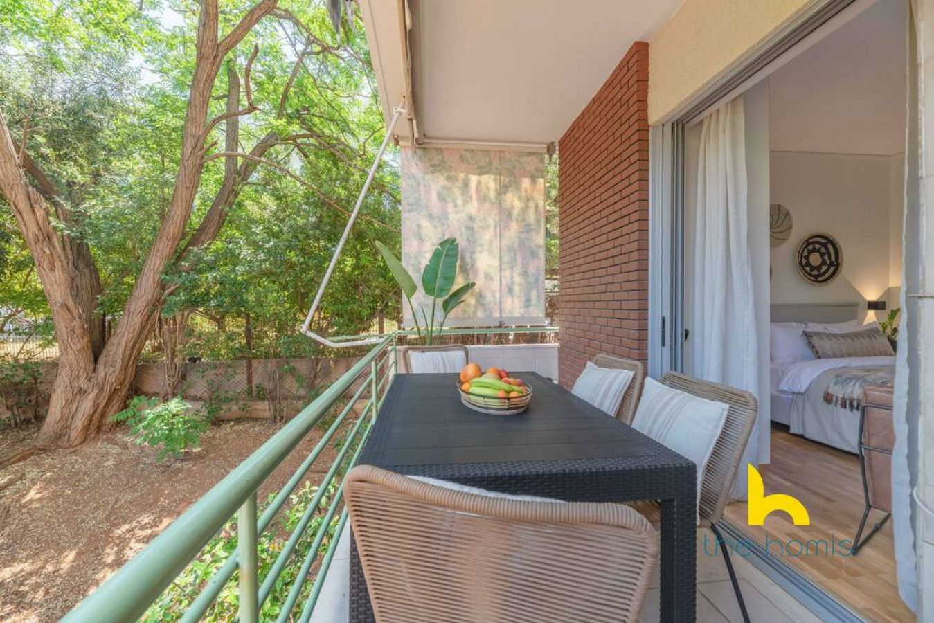 Un balcon avec une table dressée pour le petit-déjeuner, une chaise en osier et une vue sur un jardin avec des arbres et un mur de briques.