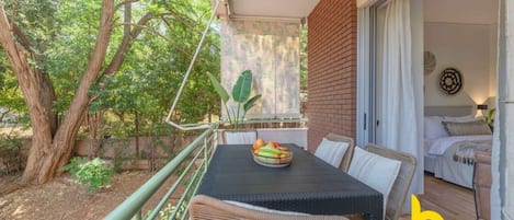 Un balcó amb una taula preparada per esmorzar, una cadira de vimet i vista a un jardí amb arbres i una paret de maó.