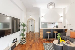 Ein modernes Wohnzimmer mit Flachbildfernseher, einer Couch, einem Couchtisch und einer Küchenzeile mit einer Theke und Hockern.