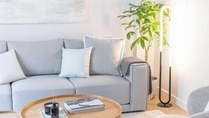 Ein modernes Wohnzimmer mit einem grauen Sofa, einem runden Couchtisch mit einem Buch und einer Kerze sowie einer hohen Stehlampe.