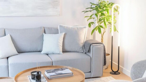 Uma sala de estar moderna com um sofá cinzento, uma mesa de centro redonda com livros e velas, um abajur e um vaso de planta.
