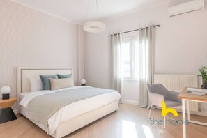 A modern bedroom with a bed, bedside tables, a desk, a chair, a window with curtains, and an air conditioning unit. - Spacious 3BR Retreat w/ 2 Baths + Fast Wi-Fi (Vironas)