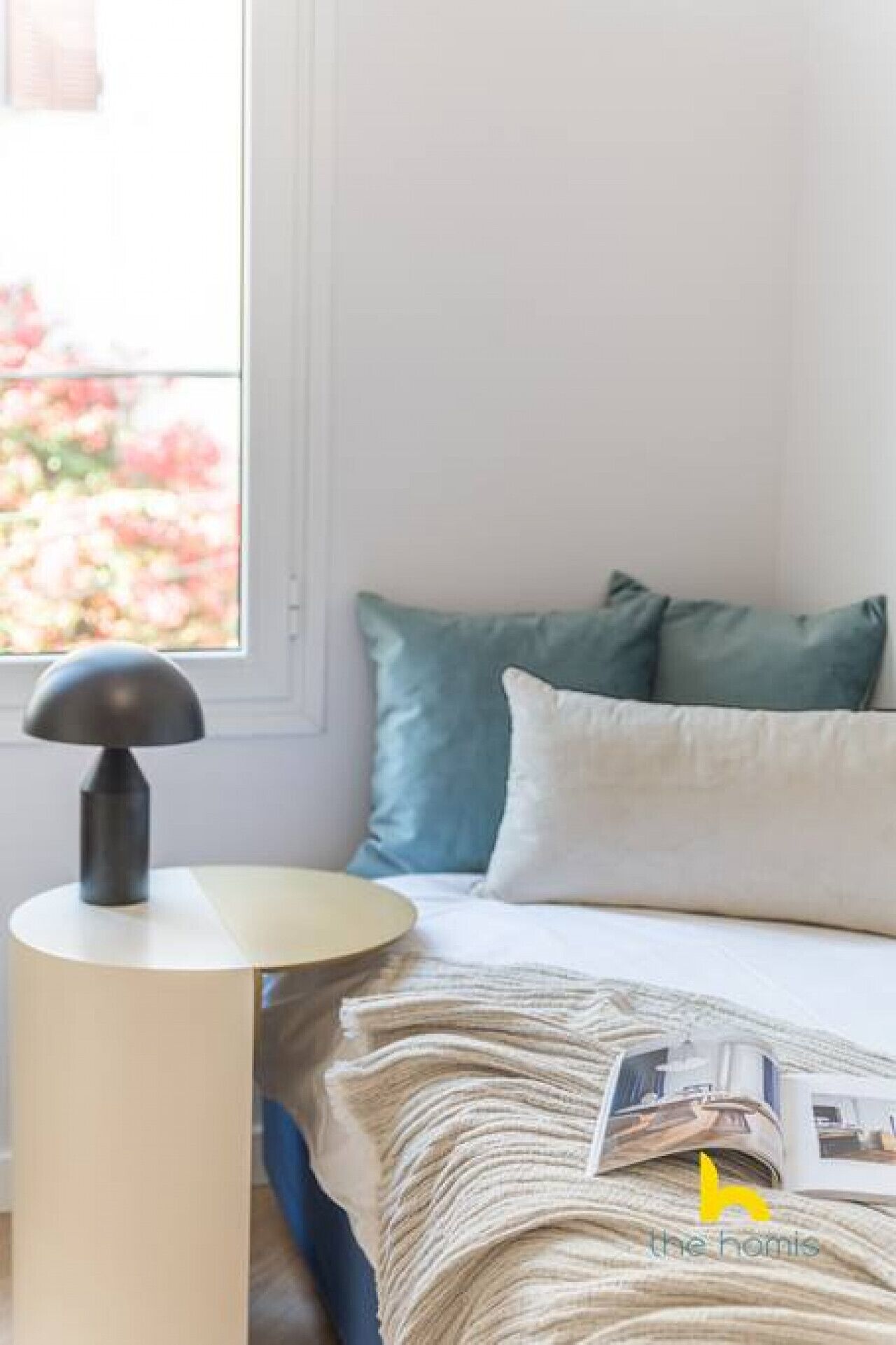 A neatly made bed with a white and teal pillow set, a small round table with a lamp, and a magazine on the bed.