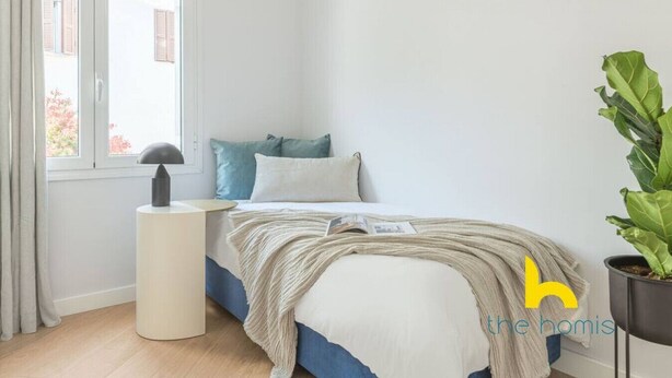 Ein Schlafzimmer mit einem Bett, einem Nachttisch mit Lampe, einem Fenster und einer Zimmerpflanze.