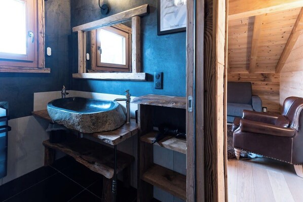 A rustic bathroom with a stone sink, wooden shelves, and a leather chair.