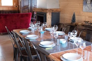 A dining table set with plates, glasses, and cutlery, surrounded by chairs in a room with a fireplace and a red chair in the background.